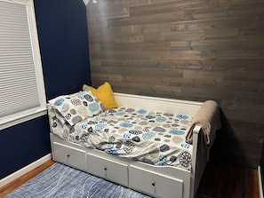 A small bed with a patterned bedspread, a single pillow, and a bedside table with drawers.
