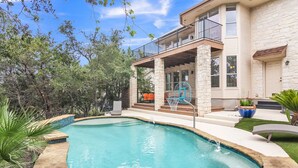 Une piscine bordée de pierres, un patio couvert avec des meubles d’extérieur et une maison en pierre dotée d’un balcon.