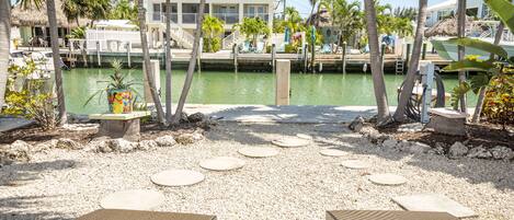 Un patio frente a la playa con mesas y sillas redondas, vista al puerto deportivo y palmeras.