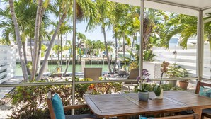 A covered outdoor seating area with a wooden table and chairs, surrounded by potted plants and overlooking a pool area with palm trees and a view of a building. - Razz's Retreat at KCB! (Key Colony Beach)