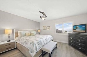 Een slaapkamer met een bed, nachtkastjes, een bankje, een televisie en een raam met zonwering.