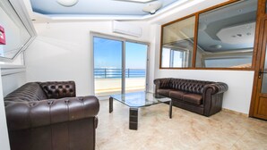 Sala de estar com dois sofás de couro, uma mesa de centro de vidro e uma grande porta de correr que dá para uma varanda com vista para o mar.