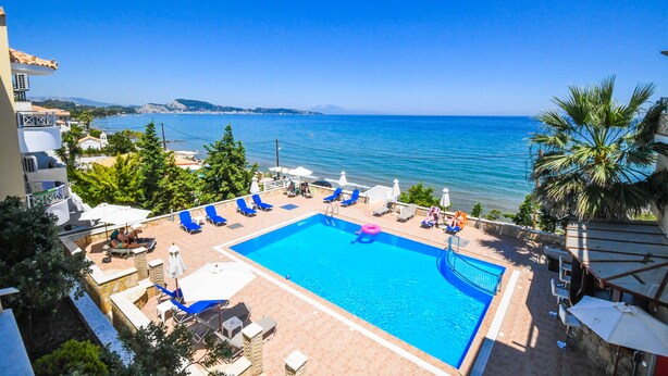 Piscina com água azul, rodeada por espreguiçadeiras e guarda-sóis, com vista para uma praia e o mar.