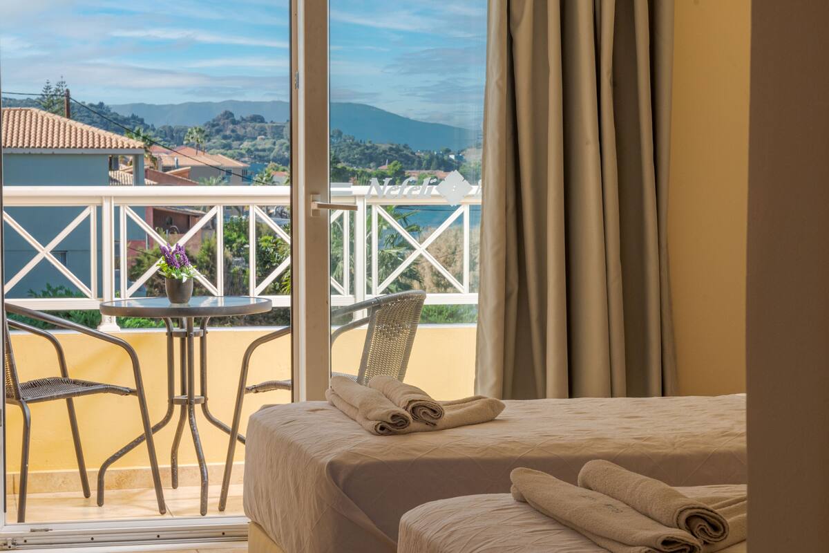 A hotel room with a balcony, a bed with neatly folded towels, a small table with a chair, and a view of the surrounding landscape.