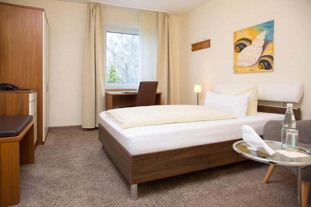 Hotelkamer met een bed, bureau, stoel, klein tafeltje met een fles en zakdoekje, en een schilderij aan de muur.
