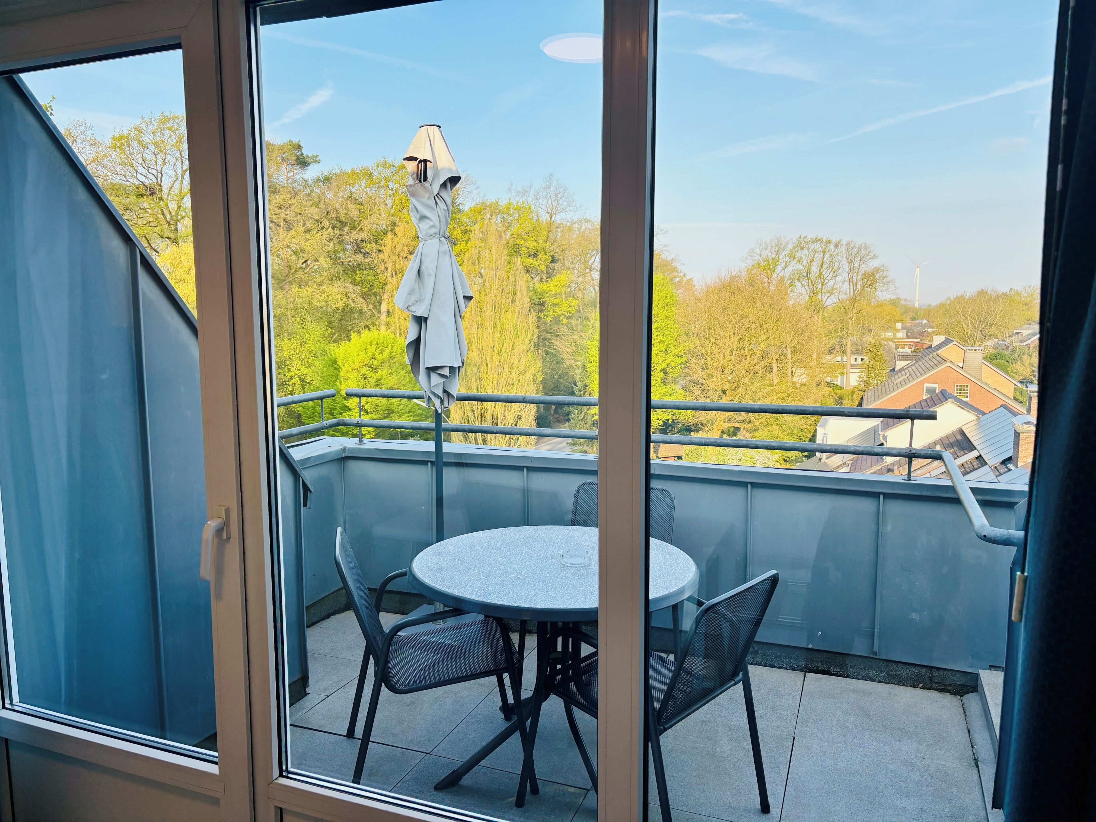 Ein Balkon mit rundem Tisch und Stühlen, einem Metallgeländer und Blick auf Bäume und Gebäude.