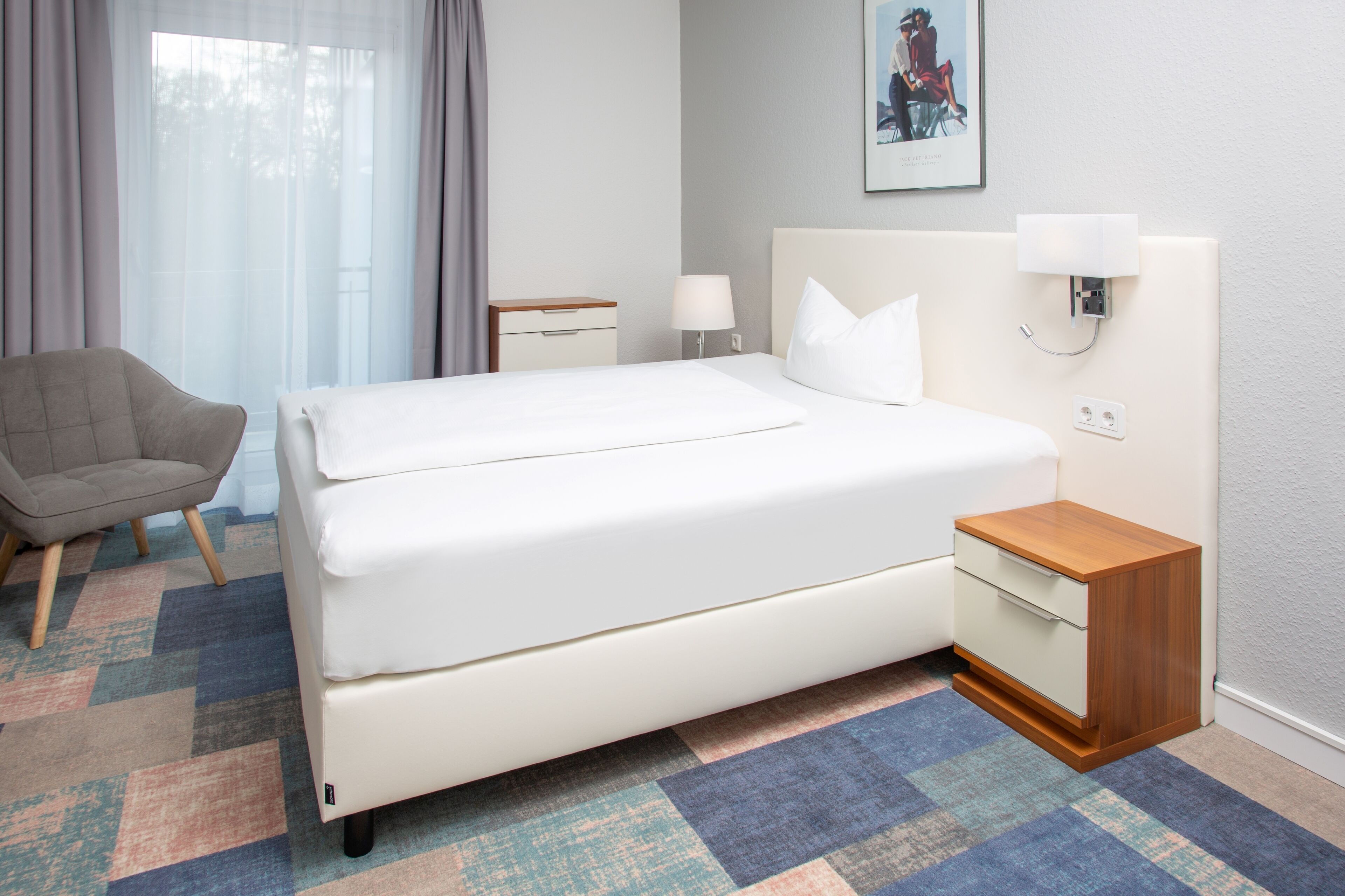 A hotel room with a bed, a chair, a bedside table, and a lamp.
