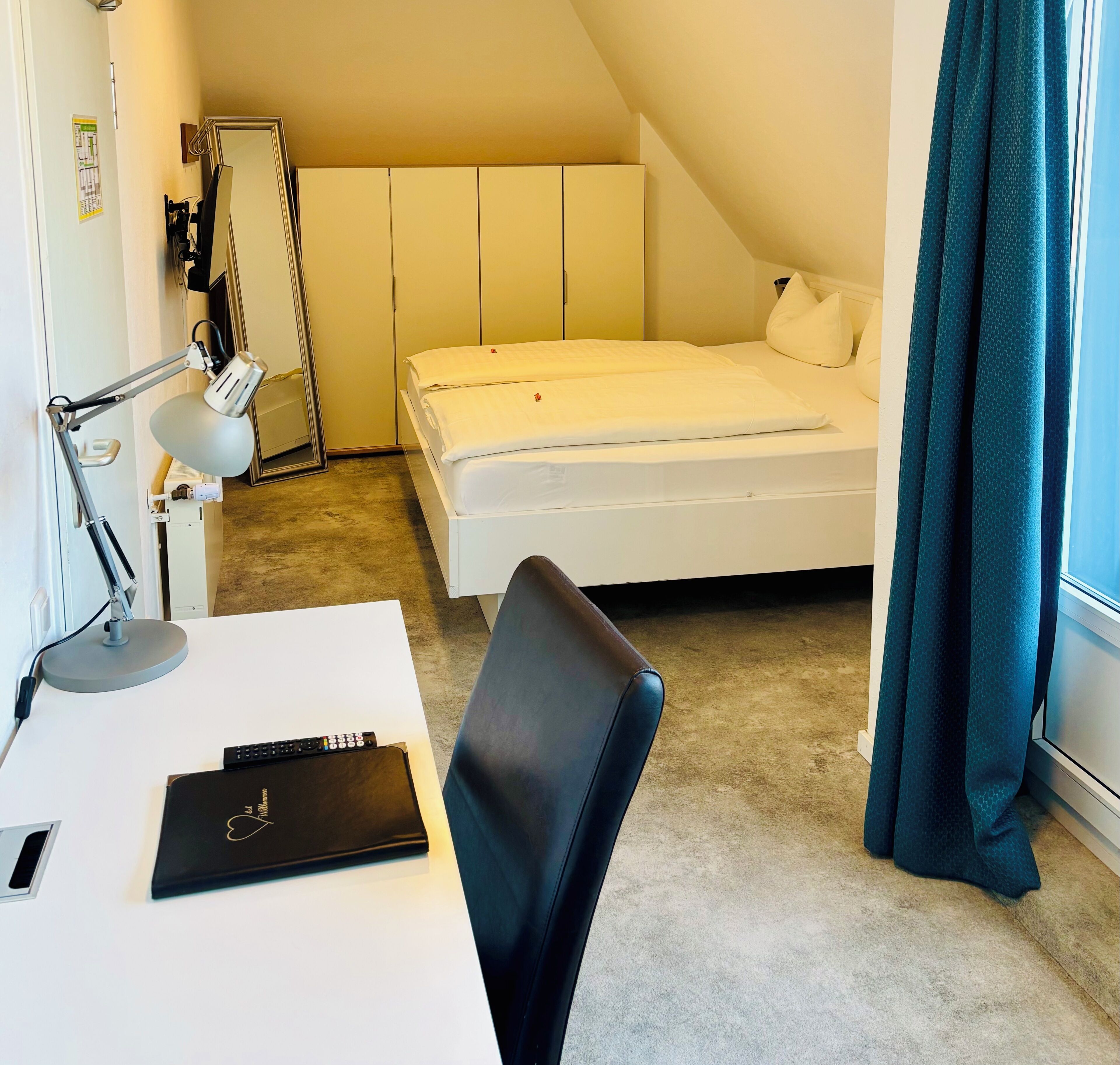 A small, neatly arranged bedroom with a single bed, a desk with a lamp, a chair, and a window with curtains.