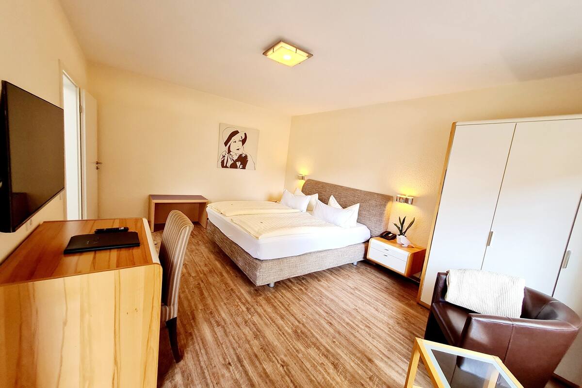 A hotel room with a bed, bedside table, lamp, wardrobe, desk, chair, and a TV.