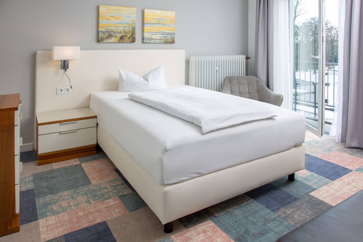 A modern hotel room with a large bed, a bedside table, a chair, and a view of the outdoors through a large window.
