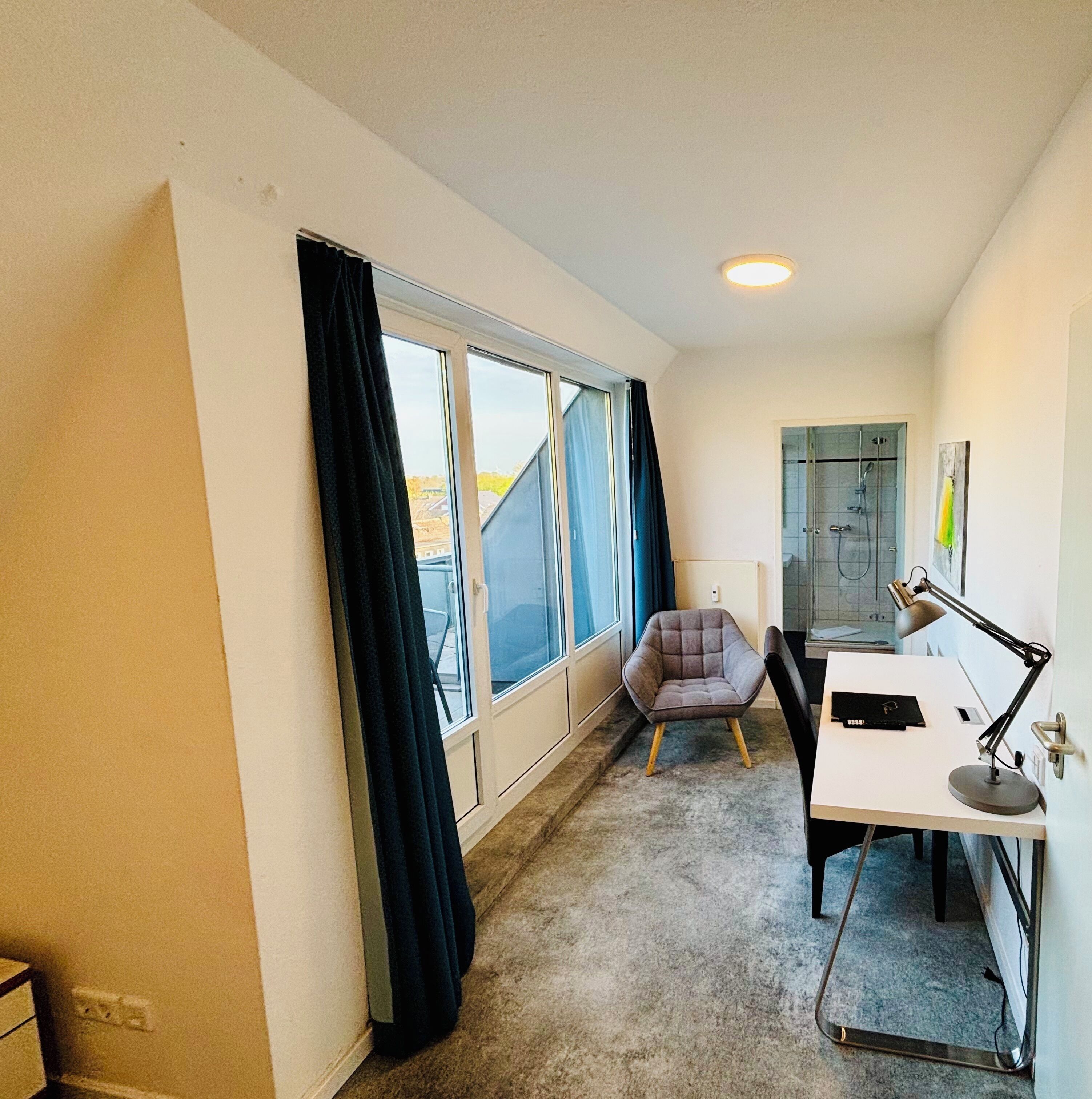 Una habitación con un ventanal grande, una silla, un escritorio con lámpara y una puerta que da a una zona de ducha.