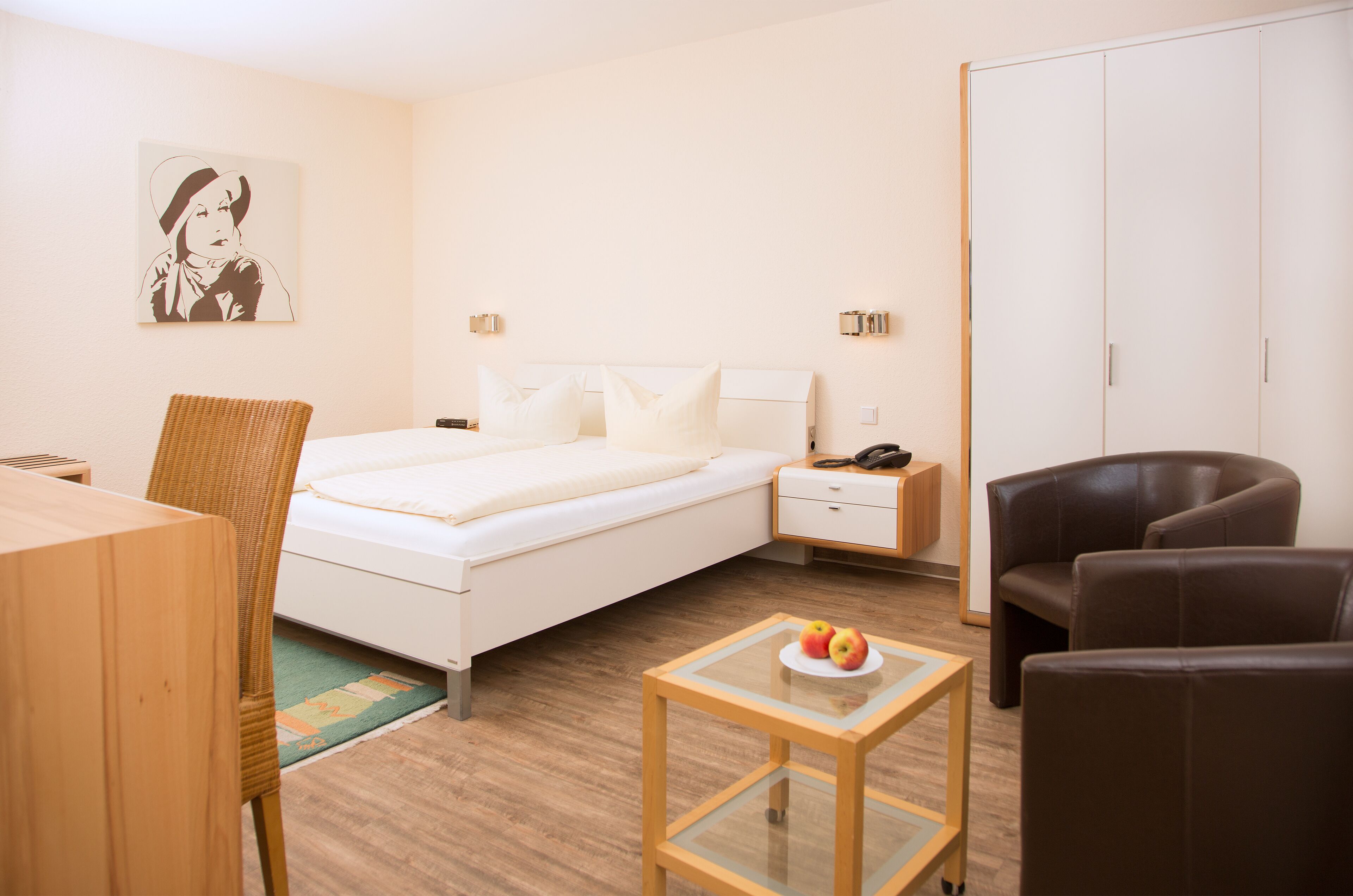 A hotel room with a bed, a chair, a small table with apples, a wardrobe, and a painting on the wall.