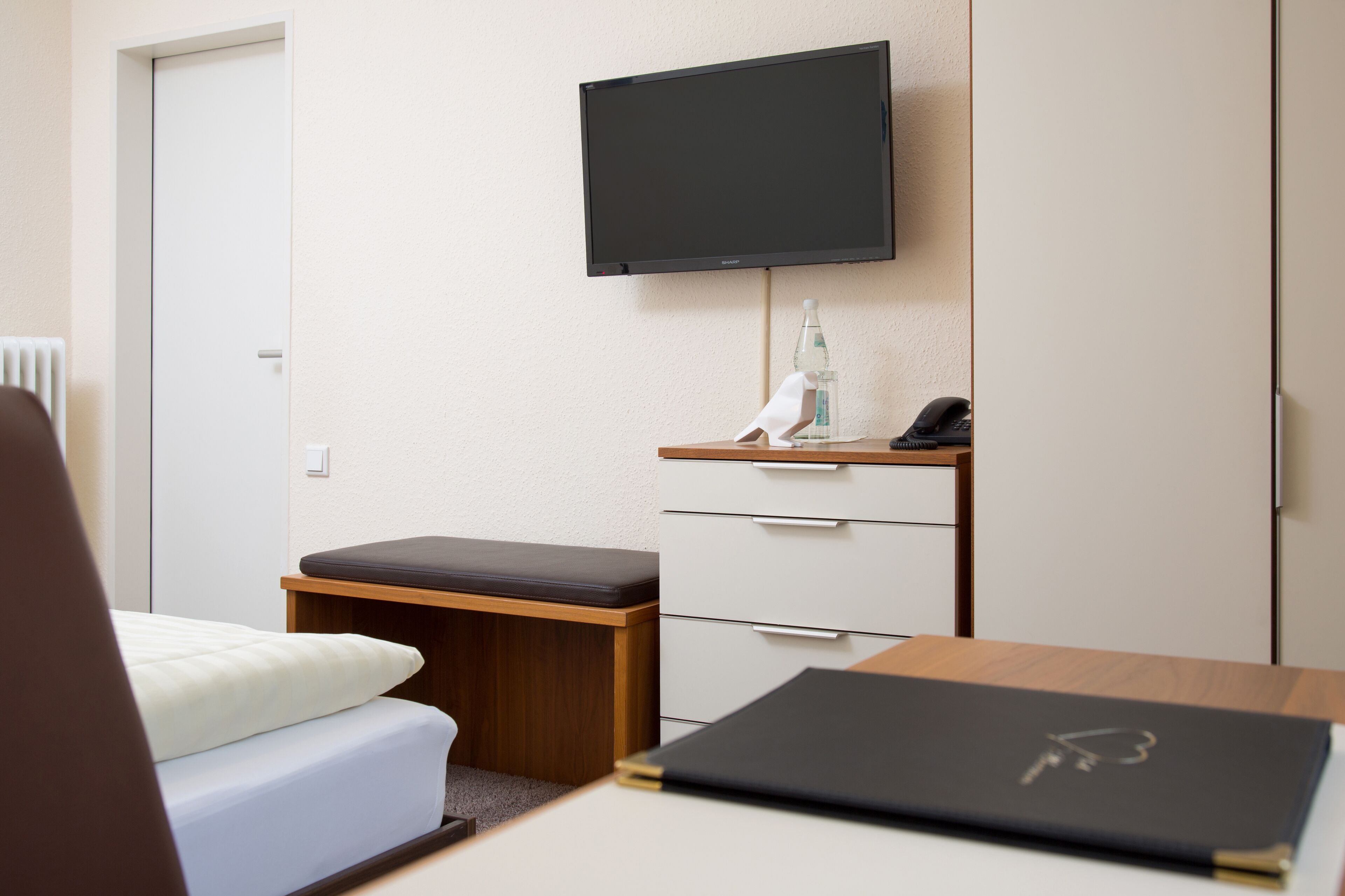 Una habitación de hotel con una cama, un escritorio con una computadora portátil, un televisor montado en la pared, una mesita con una botella y un teléfono.