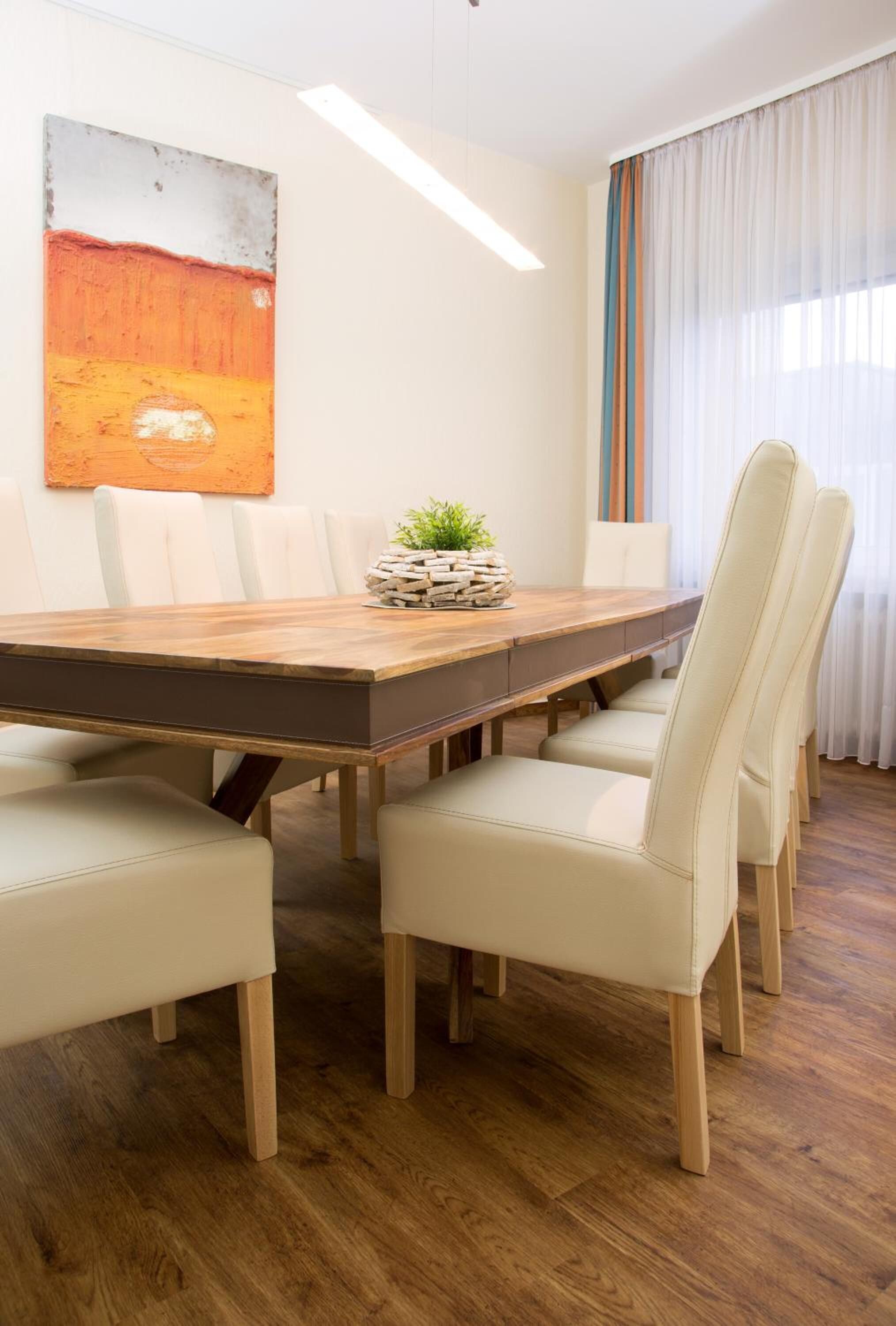 A modern dining room with a wooden table, white upholstered chairs, a potted plant, and a large abstract painting on the wall.