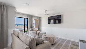 A modern living room with a large window, a ceiling fan, a flat-screen TV, and a round coffee table. - Latitude 14 - NEW OWNER! NEW LOOK! Spectacular ocean views! 1BD/2BA. Sleeps 6 (Panama City Beach)