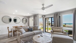 A modern living room with a sofa, armchair, and round coffee table. There are three wall-mounted mirrors and a ceiling fan. The room has large windows with a view of the beach and pool. - Latitude 14 - NEW OWNER! NEW LOOK! Spectacular ocean views! 1BD/2BA. Sleeps 6 (Panama City Beach)