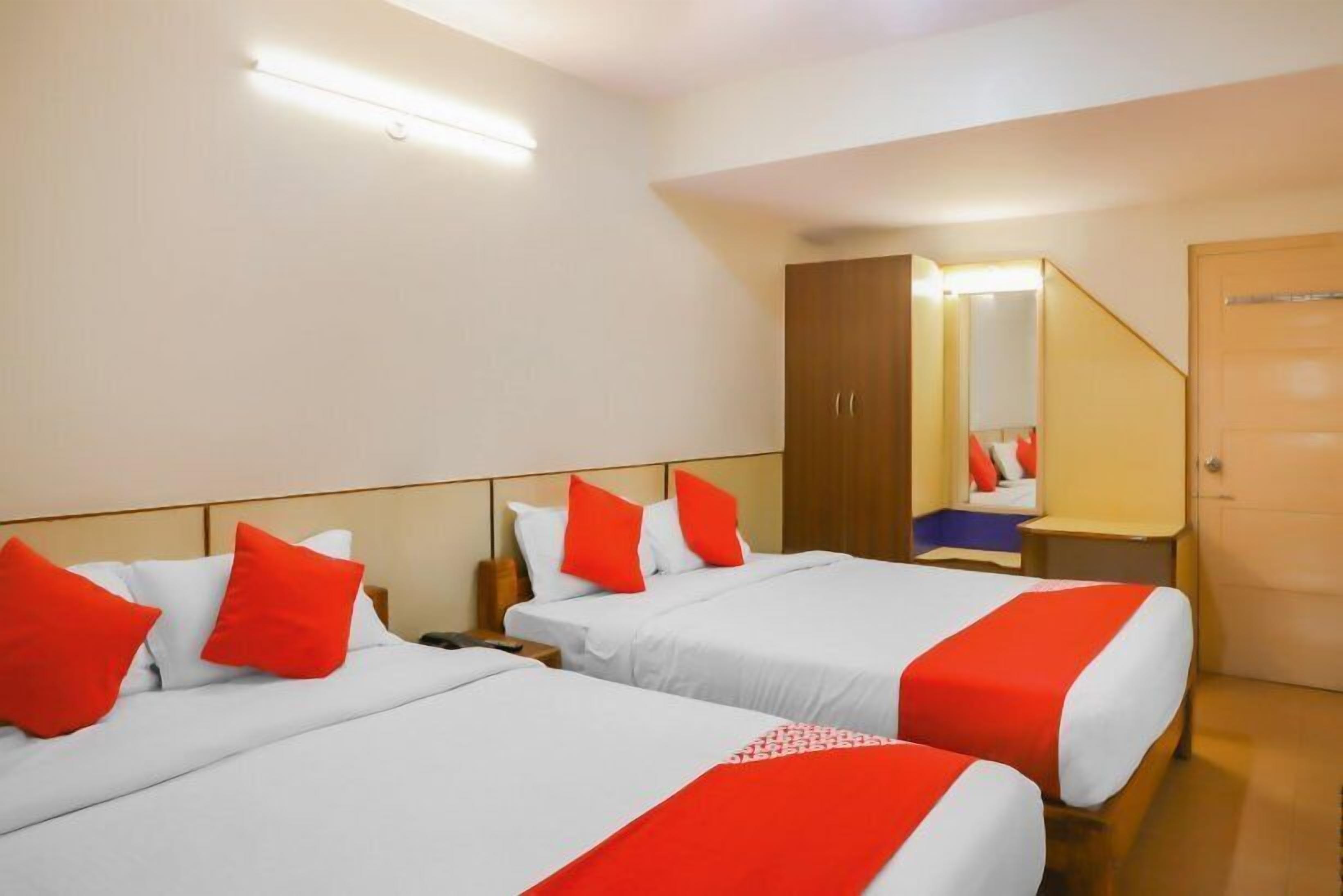 A hotel room with two beds, each with white bedding and red accents, a wooden wardrobe, a mirror, and a small bedside table.