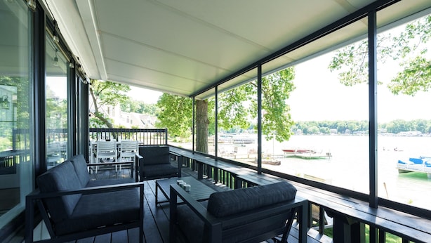 A covered outdoor area with wooden furniture and large windows overlooking a body of water.