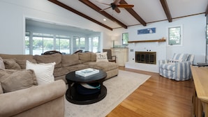 A spacious living room with a large sectional sofa, a round coffee table, and a striped armchair. There is a fireplace with a mantel, a ceiling fan, and a view of the outdoors through large windows.