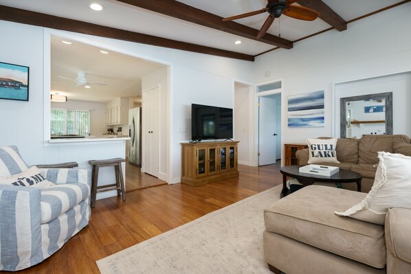 A spacious living room with a wooden floor, a beige sofa, a striped armchair, a television, and a kitchen visible in the background. - Beautiful Lakefront Gull Lake Home w/ Private Beach and Dock (Augusta)