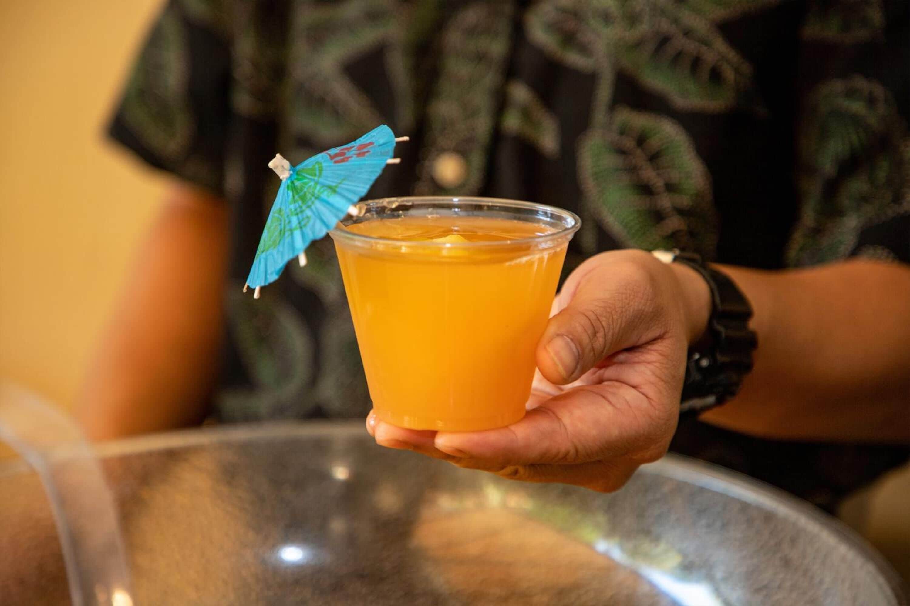 A hand holding a clear plastic cup with an orange beverage, a blue umbrella on the rim, and a watch on the wrist.