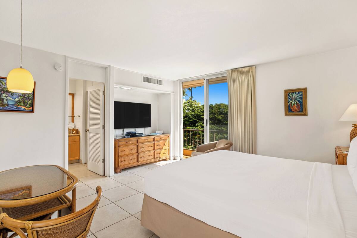 Habitación de hotel con cama, televisión, mesa de comedor con sillas, cómoda de madera y balcón con vistas a zonas verdes.