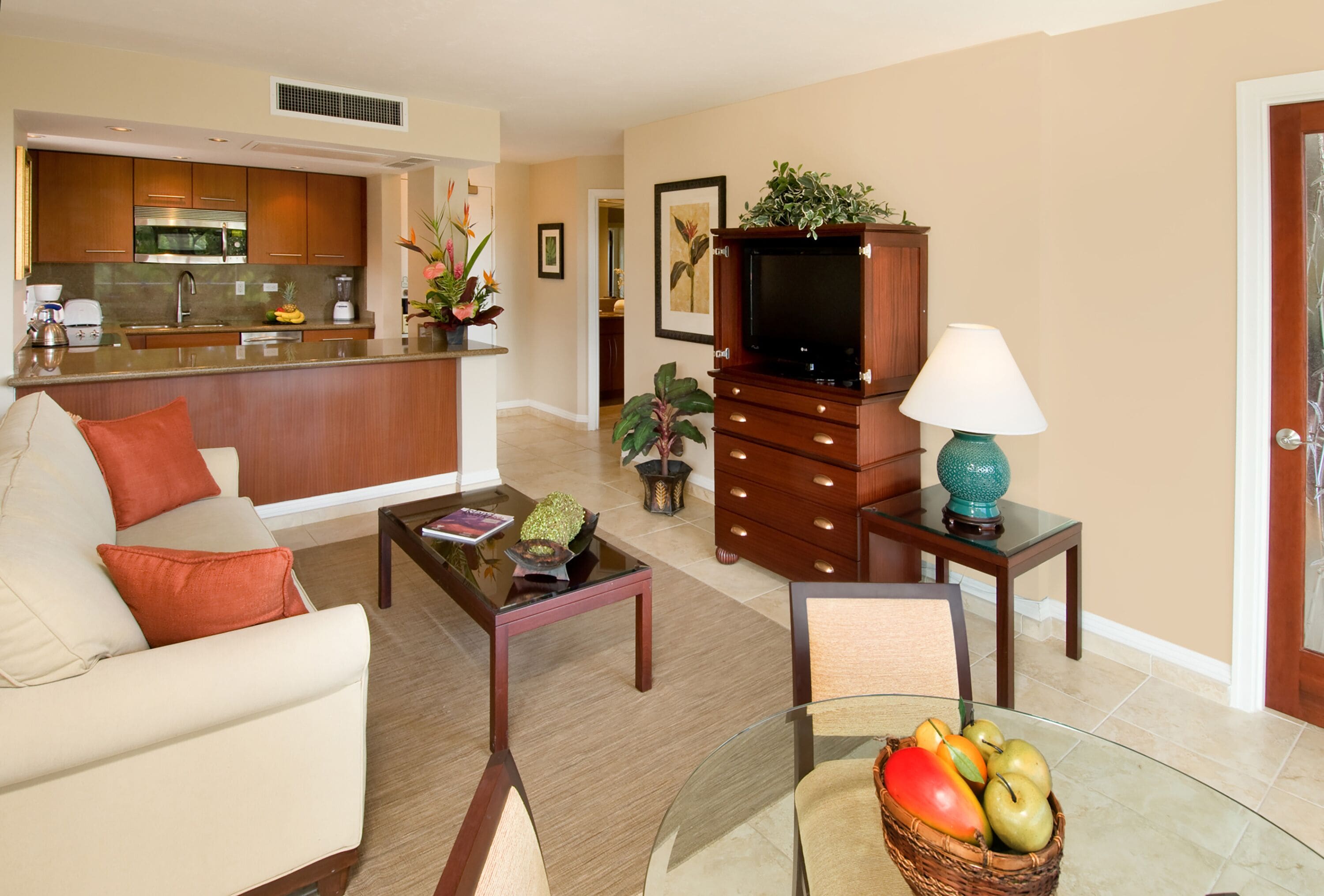 A modern living room with a sofa, coffee table, and a glass table with fruit. A kitchen is visible in the background.
