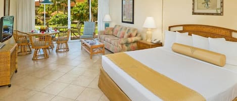 A hotel room with a large bed, a TV, a dining area with wicker furniture, and a view of the outdoors.