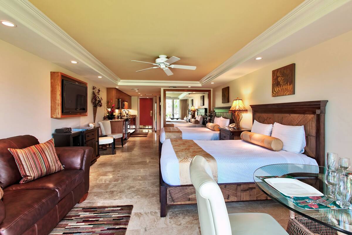 Una habitación de hotel con una cama grande, un sofá, una mesa de vidrio con una bandeja y un ventilador de techo.