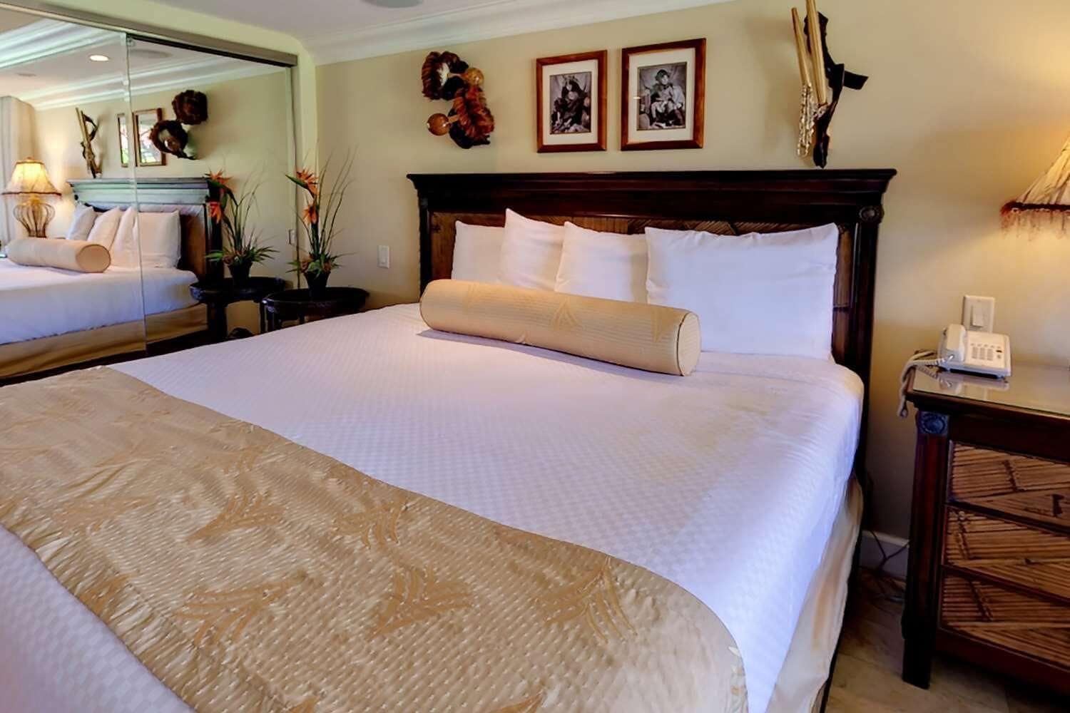 Un grand lit avec une tête de lit en bois, une literie blanche et un coussin décoratif. Des tableaux encadrés ornent le mur, une lampe se trouve sur la table de chevet et un téléphone est posé sur une petite table en bois.