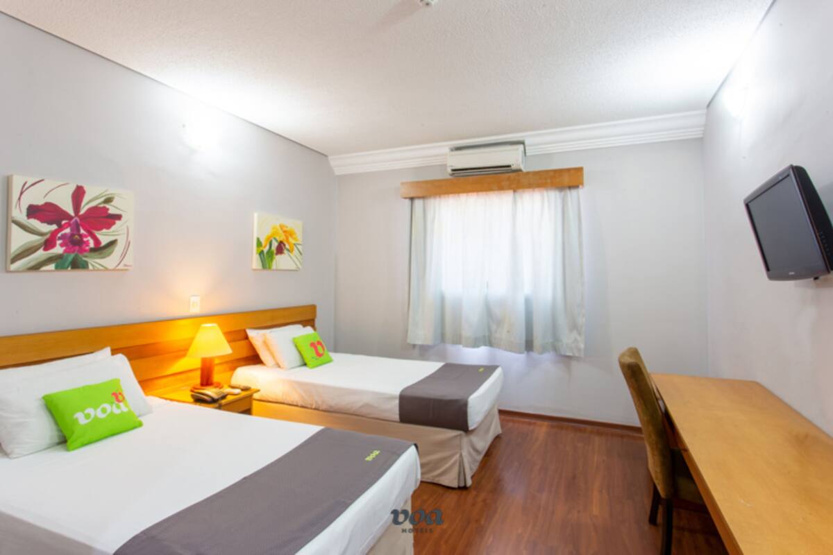 A hotel room with two beds, a desk, a chair, a TV, and artwork on the walls.
