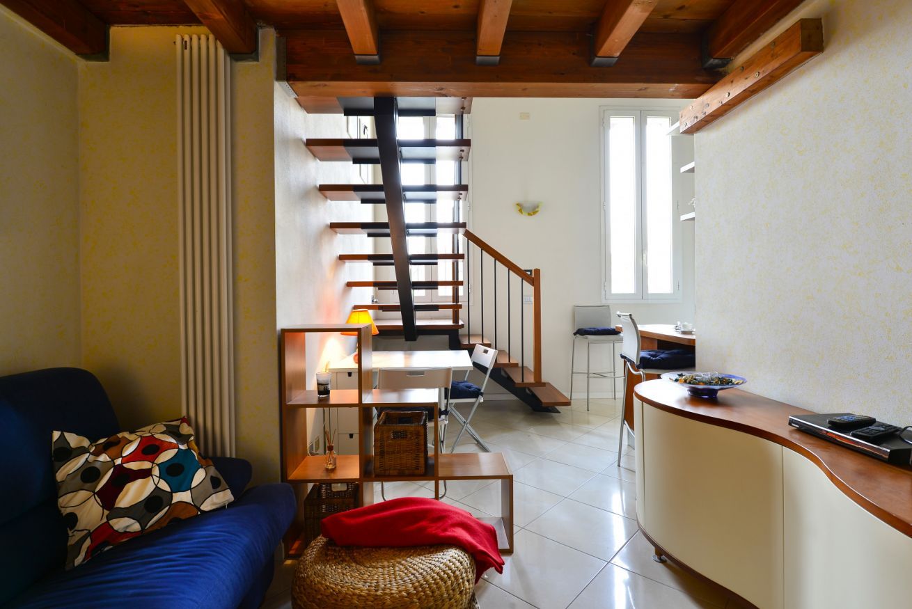 A living room with a sofa, a coffee table, a wicker ottoman, a kitchen area with a counter and chairs, and a staircase leading to an upper floor.