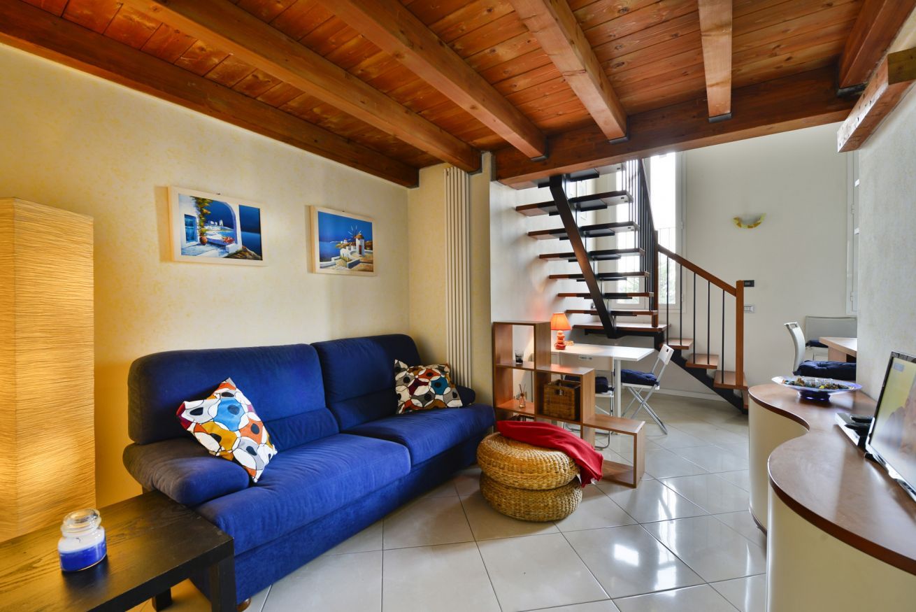 A living room with a blue sofa, a wooden ceiling, a staircase, and a small table with a lamp.