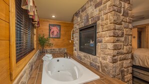 A bathroom with a stone wall, a wooden floor, a large bathtub, and a fireplace. - Beautiful Vista (Pigeon Forge)