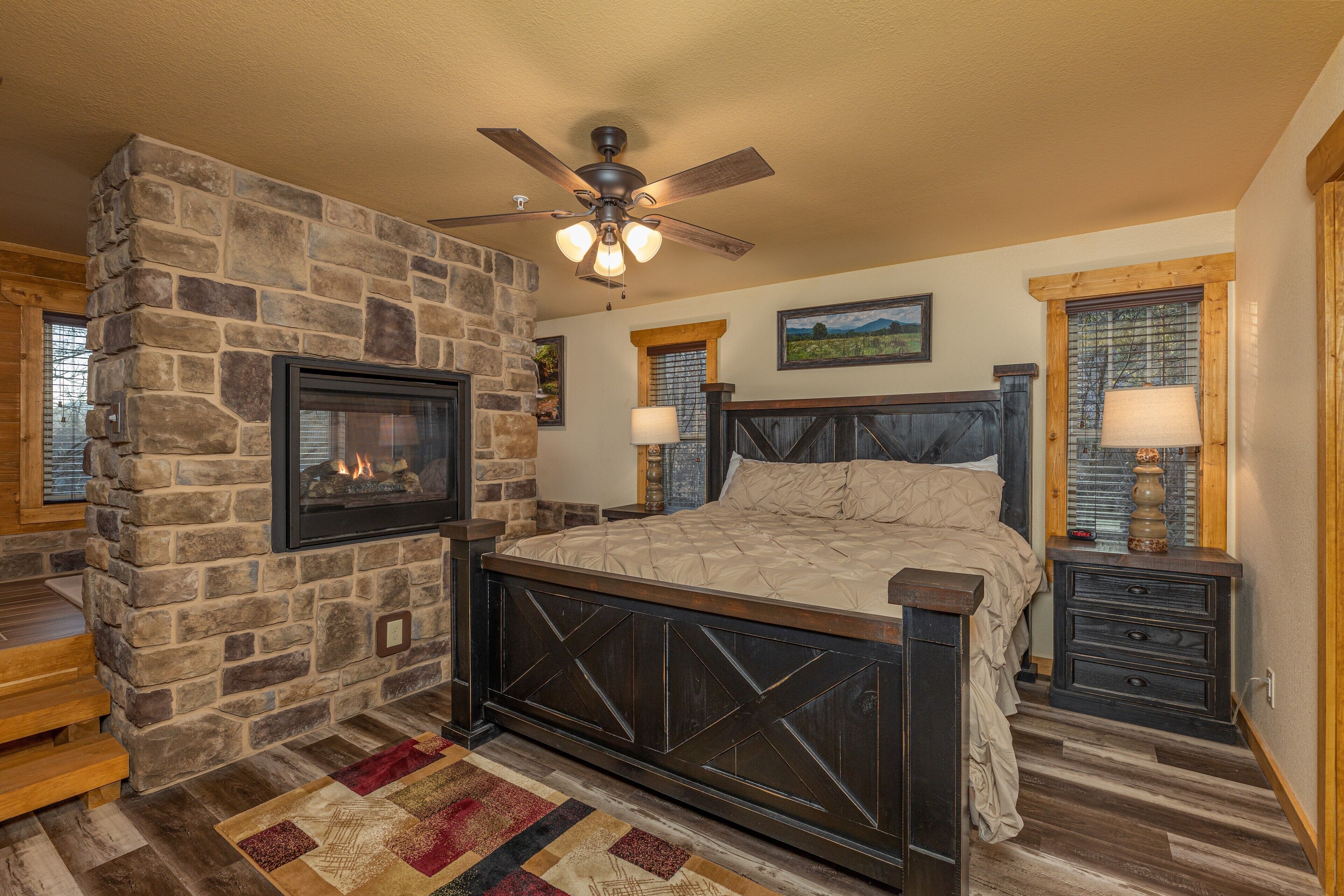 A bedroom with a stone fireplace, a bed with a headboard, a nightstand, and a window with blinds.