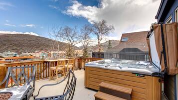 A hot tub on a deck with wooden chairs and a table, overlooking a town with buildings and hills.