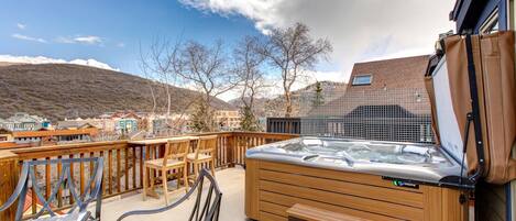 A hot tub on a deck with wooden chairs and a table, overlooking a town with buildings and hills.