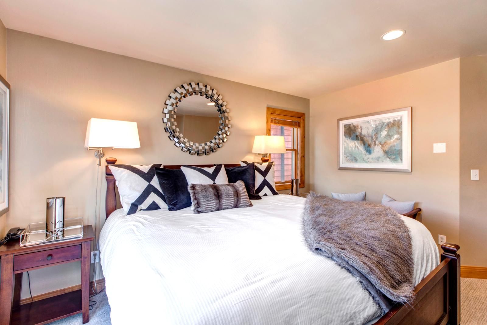 A hotel room with a large bed, two bedside tables, a mirror, a painting, and a window with curtains.