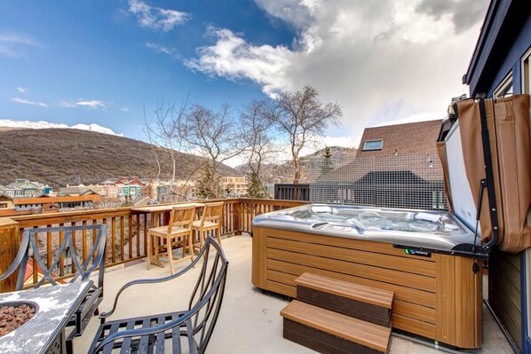 Un jacuzzi en una terraza con sillas y una mesa de madera, con vista a un pueblo con edificios y colinas.