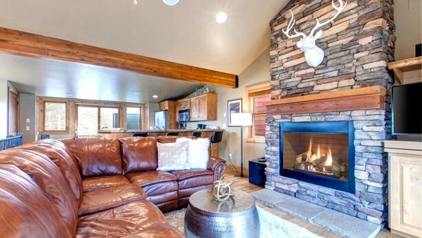 A living room with a stone fireplace, leather sofa, and a kitchen in the background. - KBM Resorts | Private Home | Split AC in living room | Walk to Town and to Ski Access, Hot Tub and Fire Pit! PNF-860 (Park City)