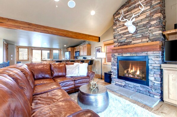 A living room with a stone fireplace, leather sofa, and a kitchen in the background.