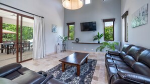A spacious living area with a leather sofa, a wooden coffee table, and a TV mounted on the wall. There is a sliding glass door leading to an outdoor patio with a table and chairs. - Casa Bonita Beach (Tamarindo)