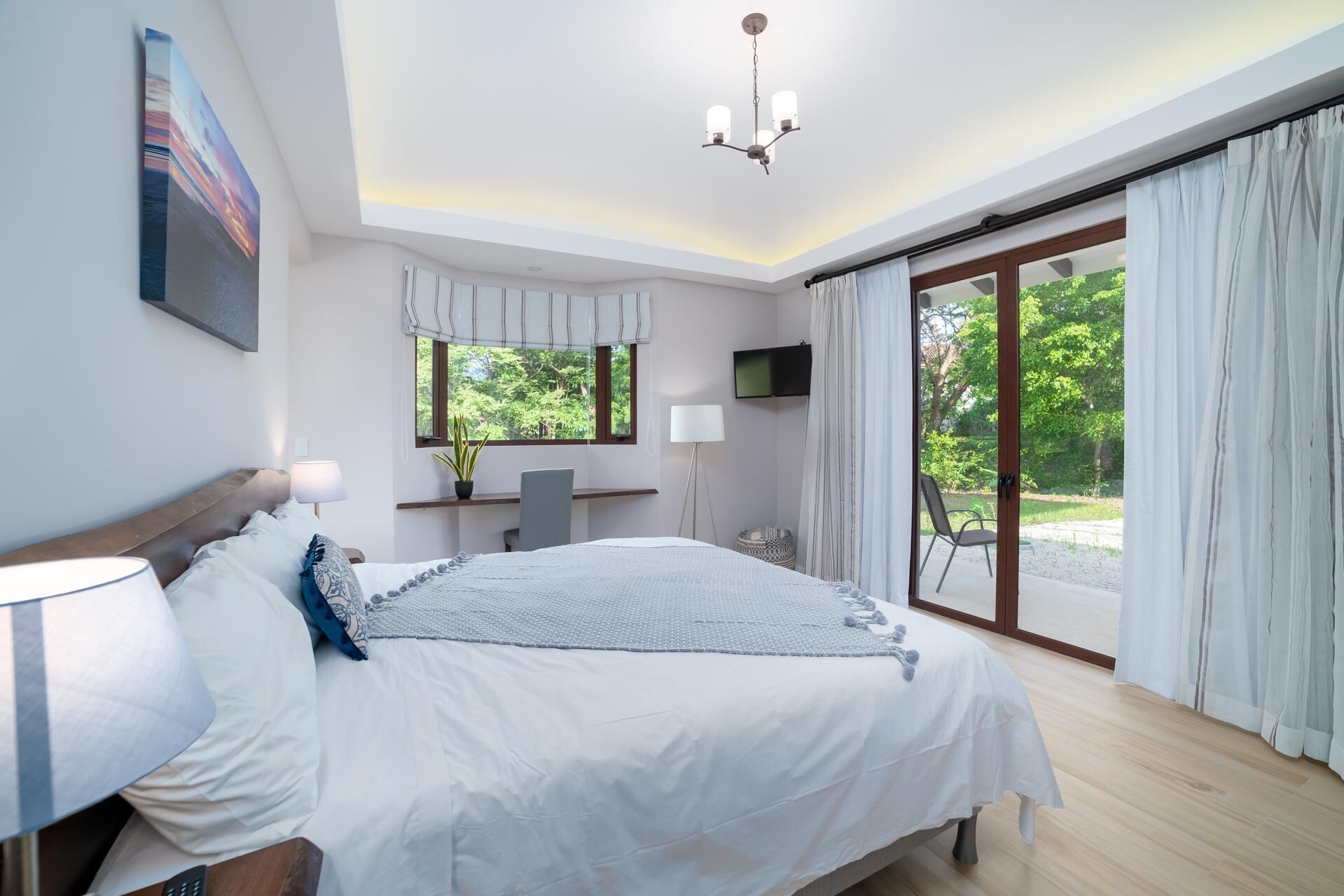 A bedroom with a bed, bedside table, lamp, TV, and a view of greenery through a sliding glass door.