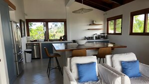 A modern kitchen with white appliances, a dining area with white chairs and blue cushions, and large windows offering views of greenery.