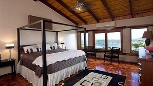 A four-poster bed with a white and brown bedspread, a wooden ceiling, and a view of the ocean through large windows.