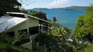 Una casa moderna con un balcón grande que da a un mar azul cristalino y montañas a lo lejos.