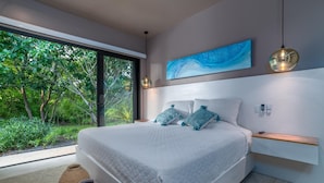 Una camera da letto moderna con un letto grande, vista sul verde attraverso una porta scorrevole in vetro e un quadro astratto blu appeso al muro.