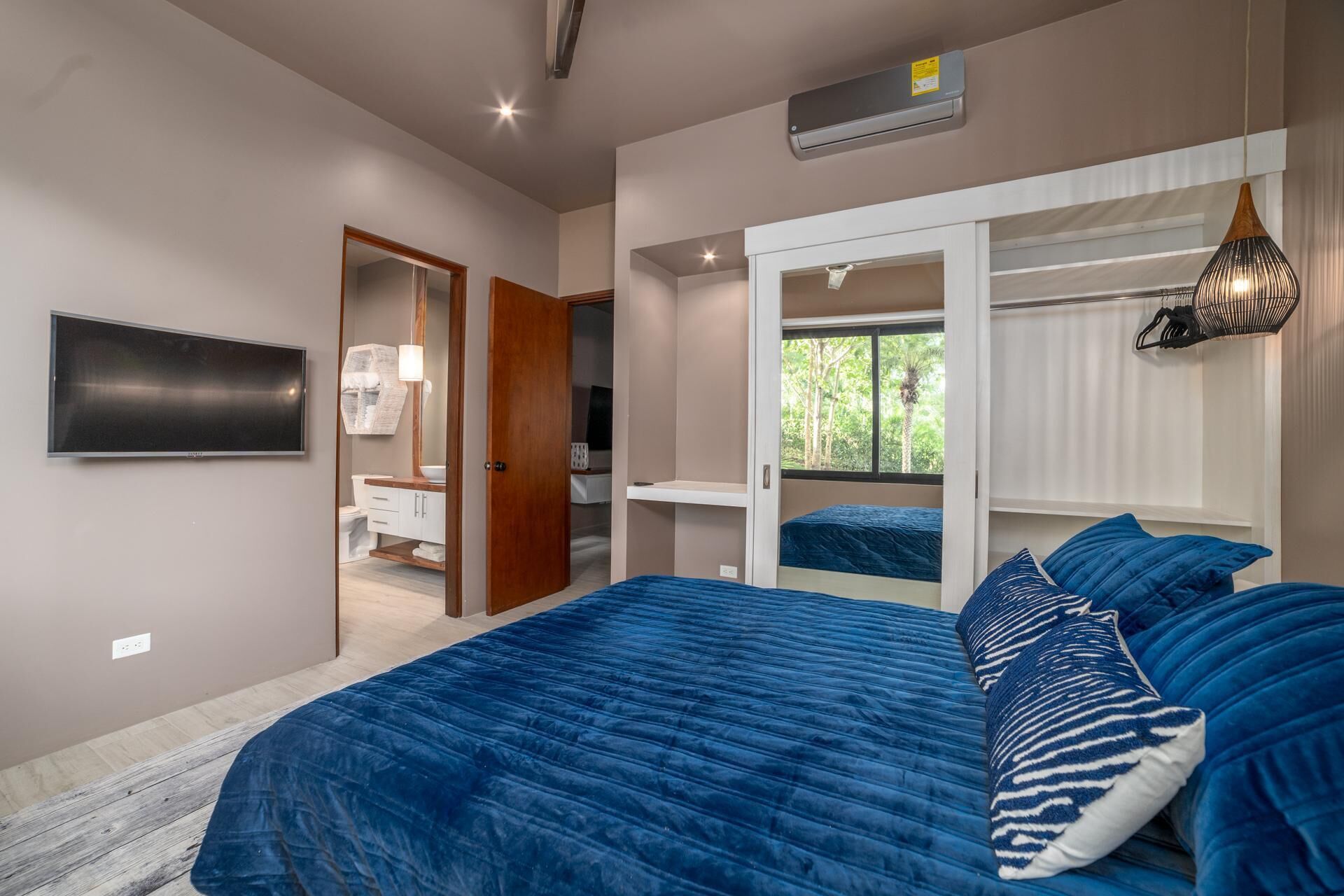 A bedroom with a large bed, a TV, a walk-in closet, and a window with a view of greenery.
