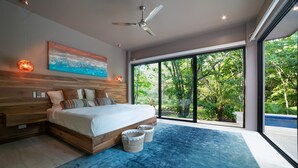 Camera moderna con un letto grande, una ventola a soffitto, un quadro e vista sul verde attraverso ampie finestre.