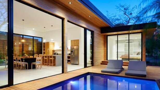 Una casa moderna con piscina, terraza de madera y puertas de cristal que dan a un comedor y cocina.